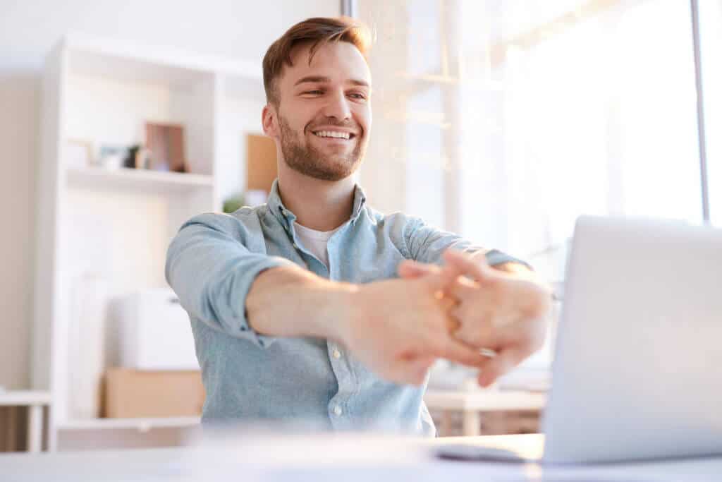Blogg.10 tips til god ergonomi.sittbedre.no. På bildet ser du en mann som sitter på kontoret sitt og smiler mens han tøyer ut fingrene mot datamaskinen.