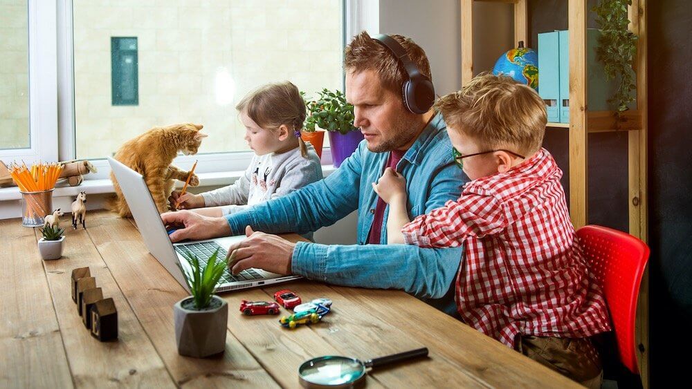 Blogg. Nye regler for hjemmekontor. Mann sitter hjemme å jobber på laptop ved kjøkkenbordet sammen med barna. Den ene sønnen henger på skuldrene for å få oppmerksomhet.